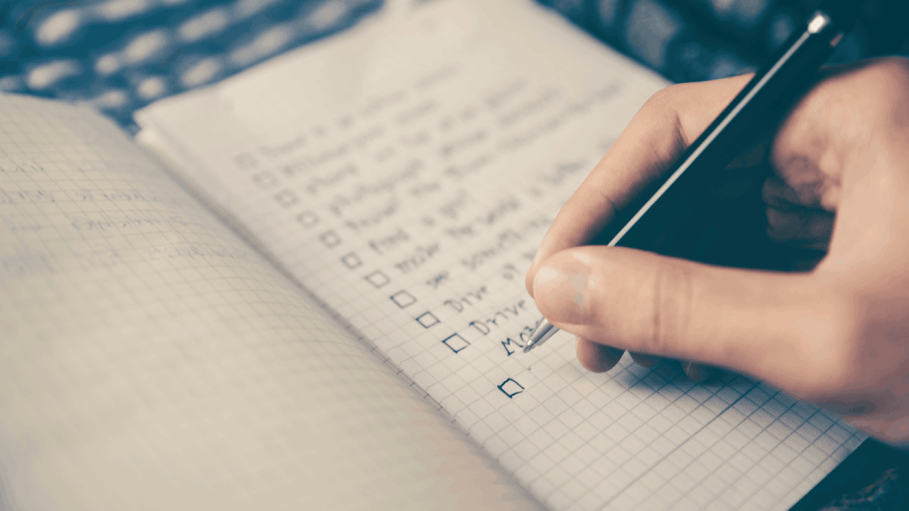 A person is writing on a checklist with a pen, surrounded by office supplies and stationery in an indoor setting.